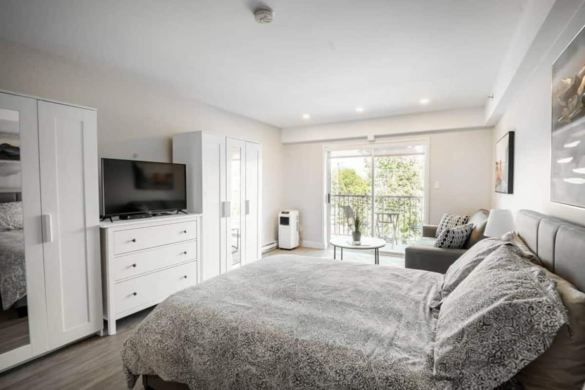 A modern bedroom with a large bed, a TV, a wardrobe, and a balcony with a view.