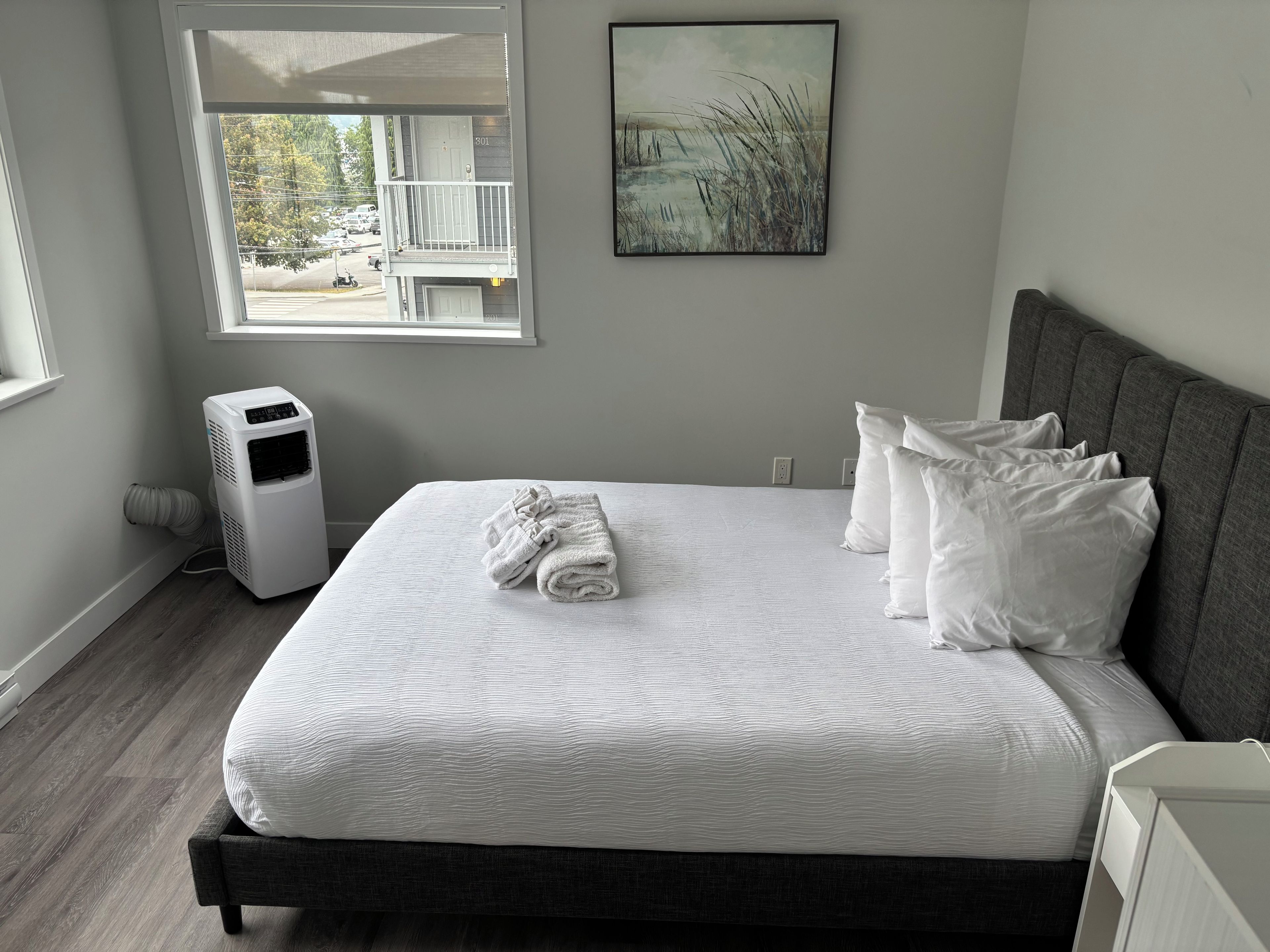 A bedroom with a bed, white pillows, a white blanket, a gray headboard, a window with a view of a building and trees, and a wall-mounted picture.