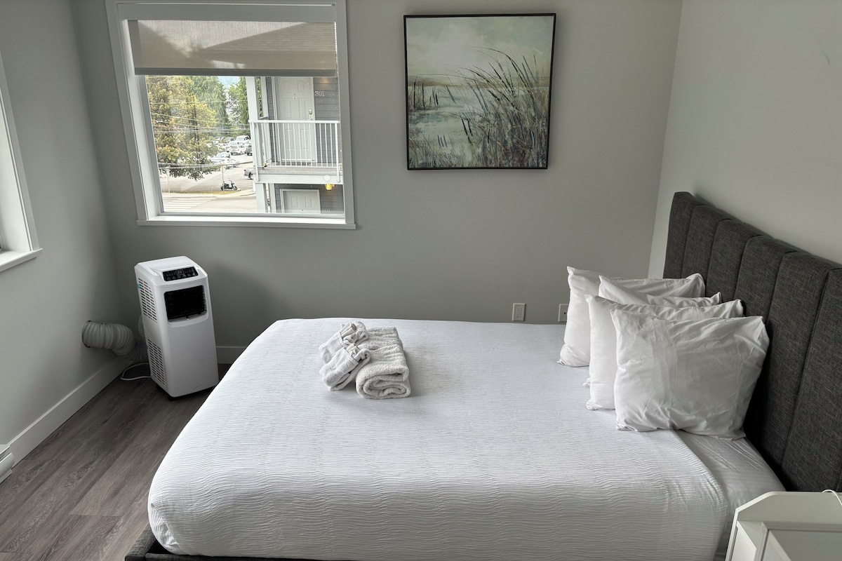 A bedroom with a bed, white pillows, a white blanket, a gray headboard, a window with a view of a building and trees, and a wall-mounted picture.