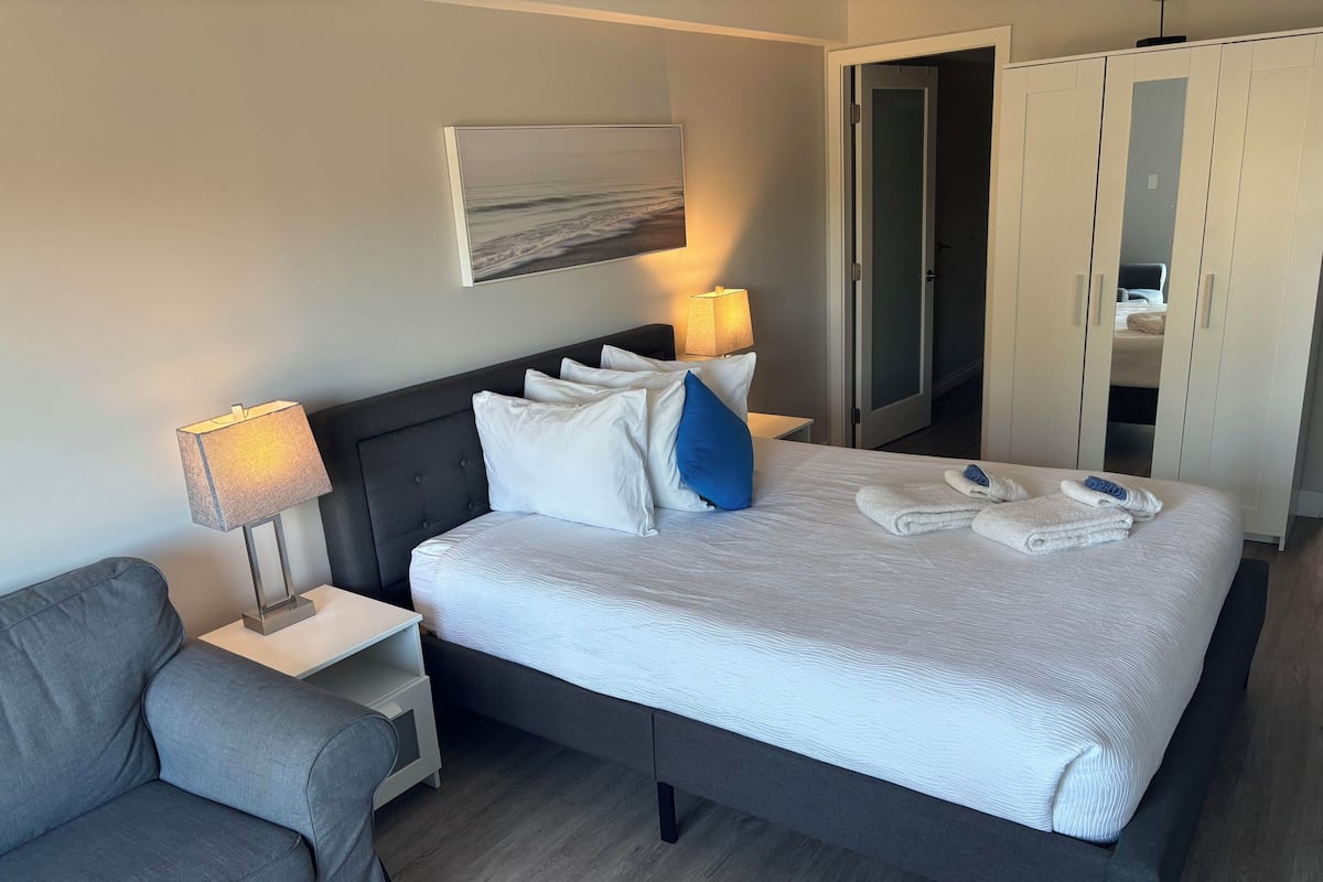 A neatly made bed with pillows, a bedside lamp, a grey armchair, and a wardrobe.