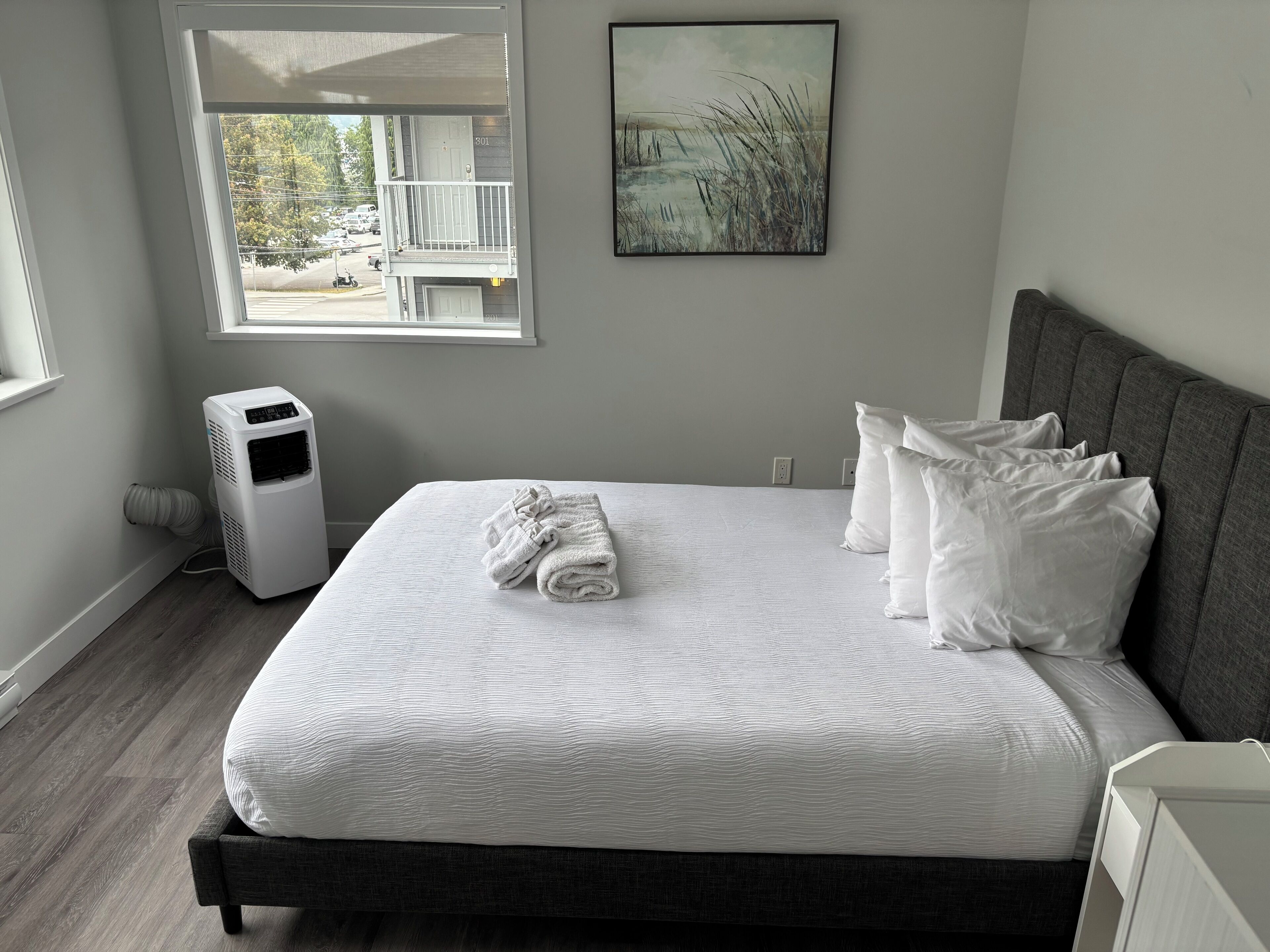 Un dormitorio con cama, almohadas blancas, una colcha blanca, cabecera gris, ventana con vista a un edificio y árboles, y un cuadro colgado en la pared.