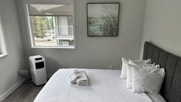 A bedroom with a bed, white pillows, a white blanket, a gray headboard, a window with a view of a building and trees, and a wall-mounted picture.