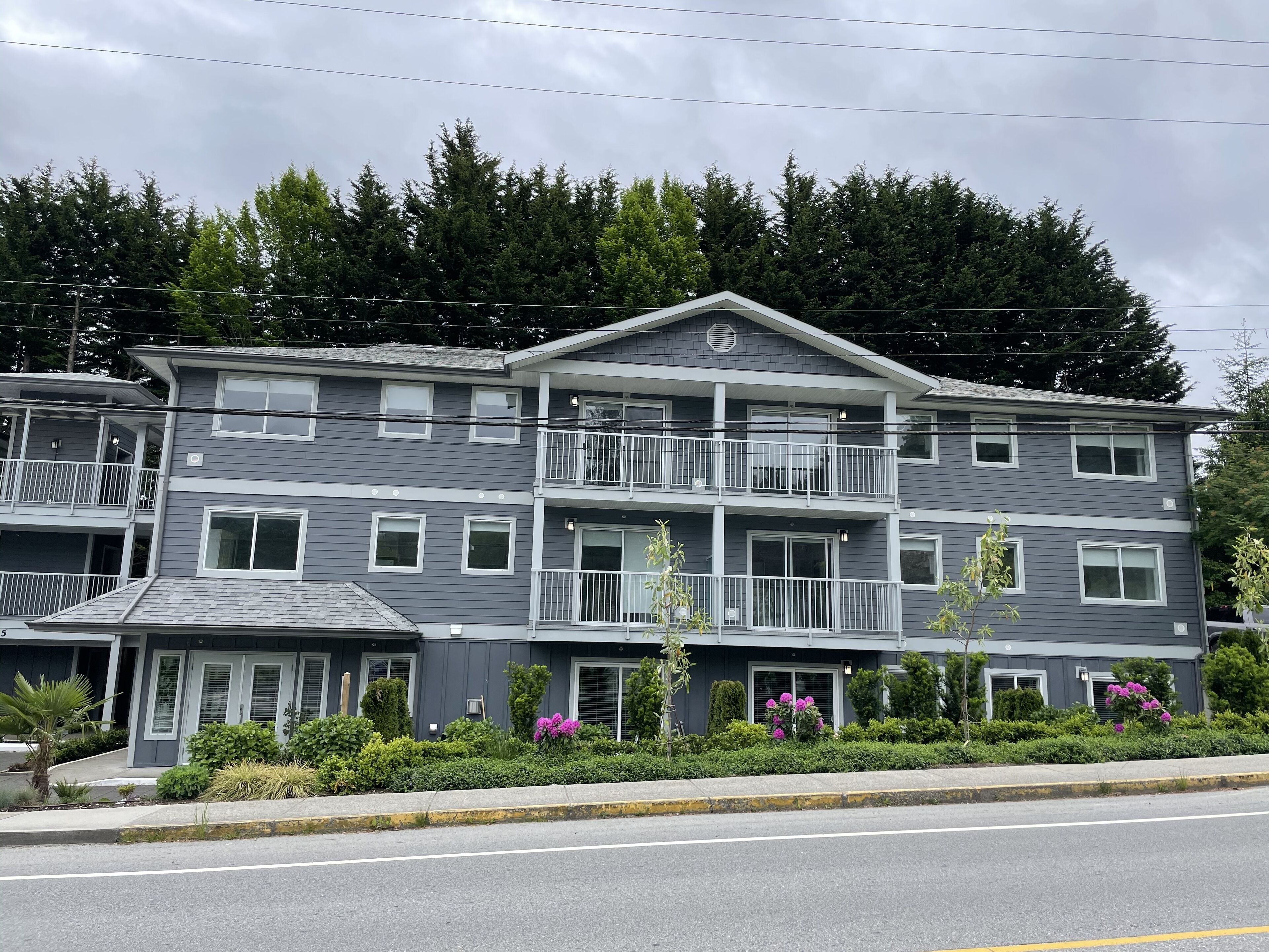 A multi-story hotel with balconies, a central entrance, and a landscaped garden with shrubs and flowers.