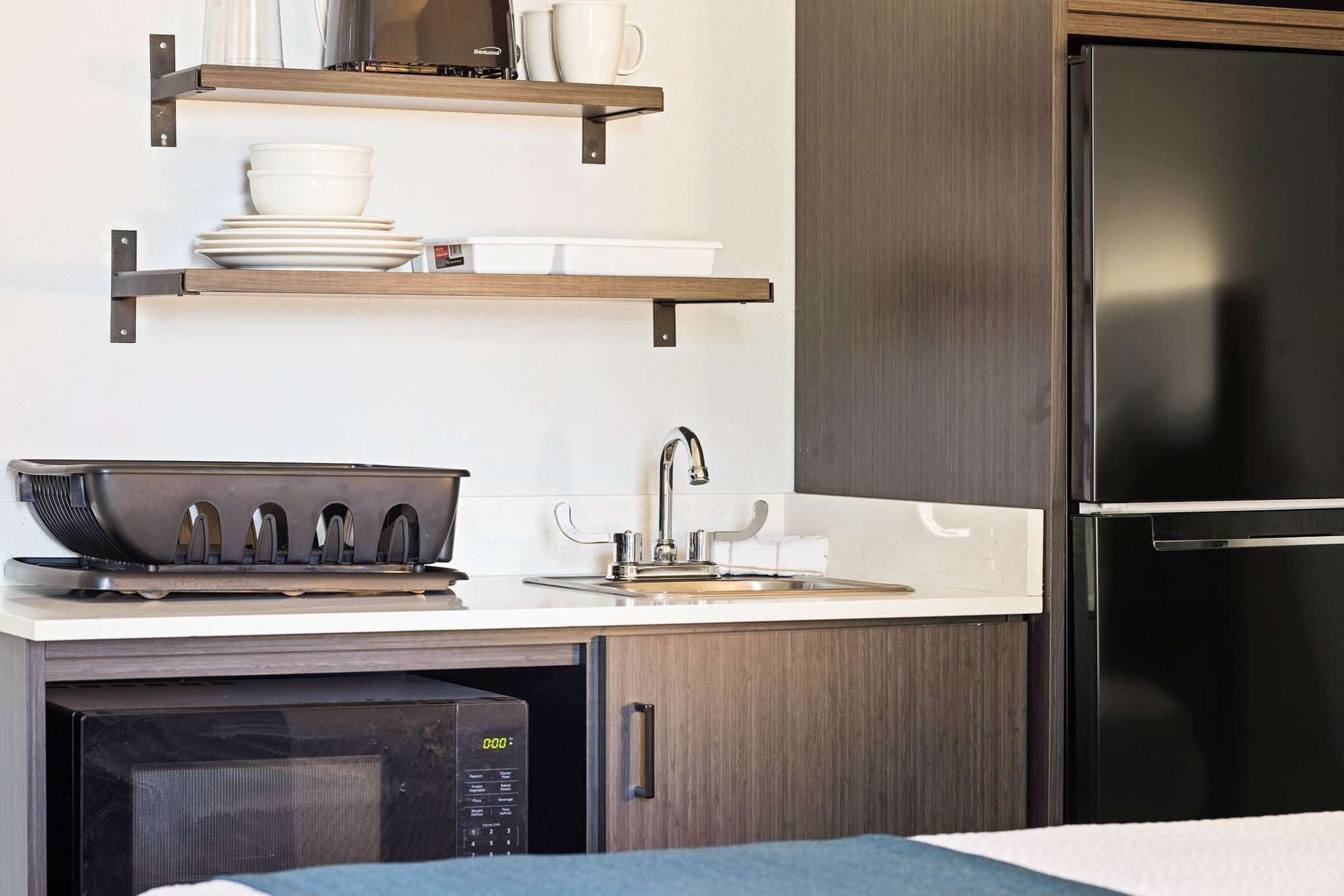 A compact kitchen with a microwave, stove, and stainless steel refrigerator. There are wooden shelves above the counter holding dishes and a coffee maker.