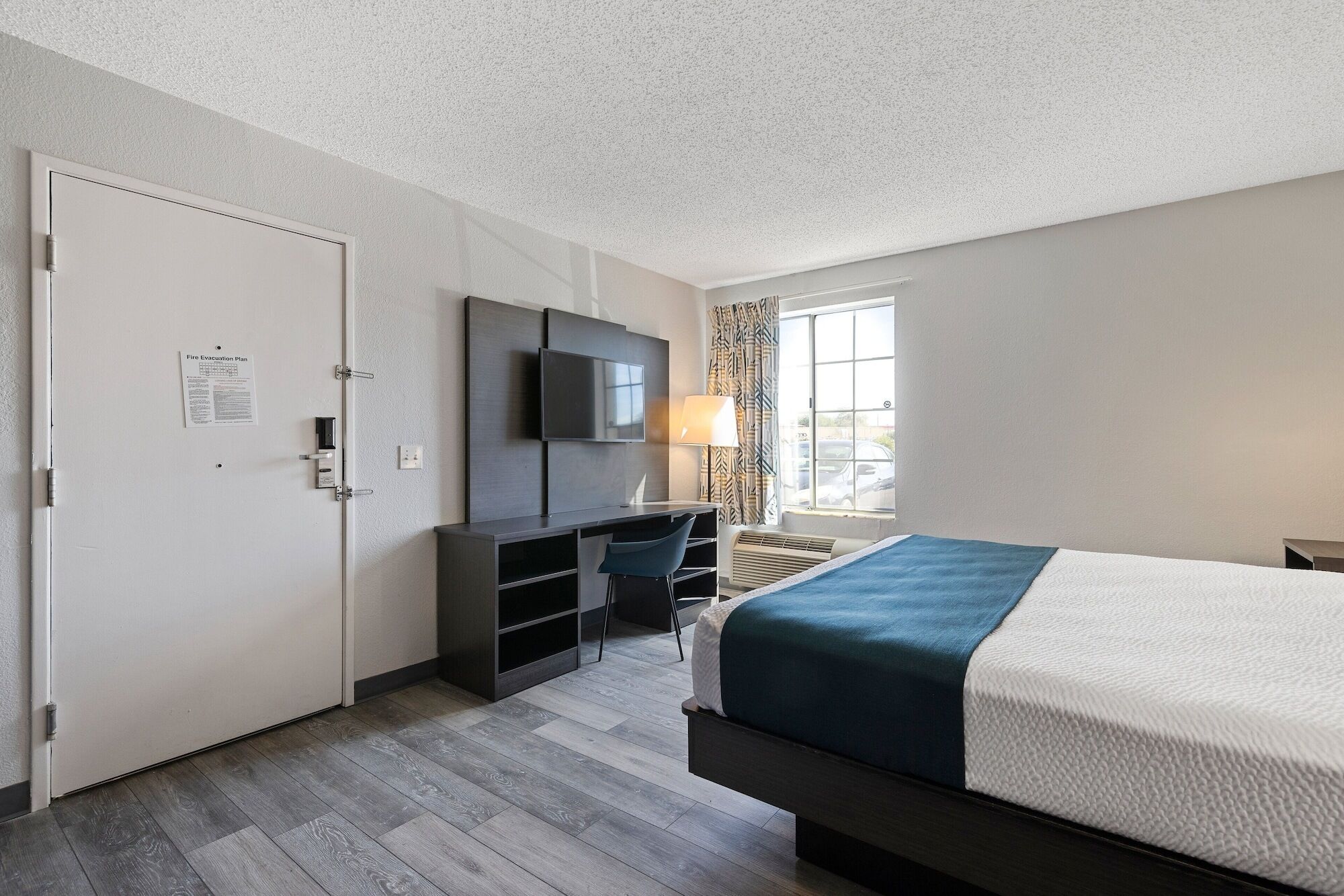 A hotel room with a bed, a desk with a TV, a window with curtains, and a fire evacuation plan on the door.