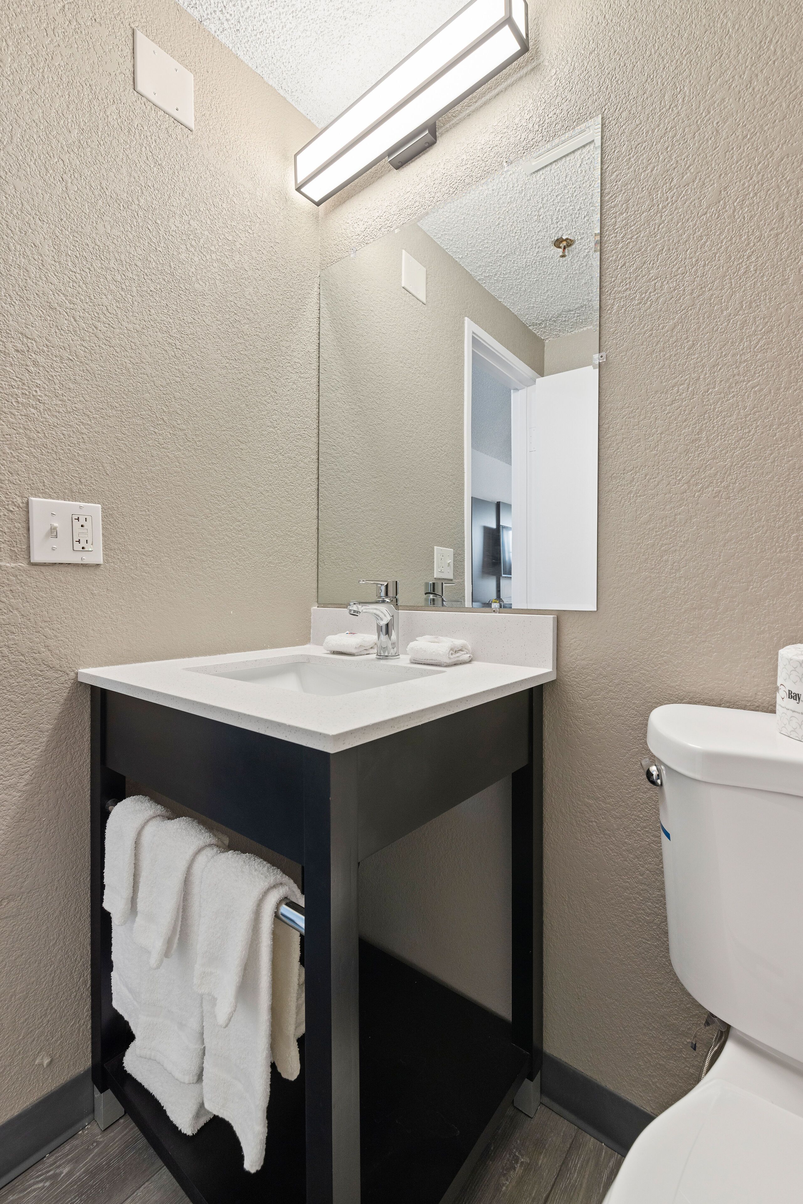 Un baño moderno con un lavamanos blanco, tocador oscuro y un inodoro blanco.