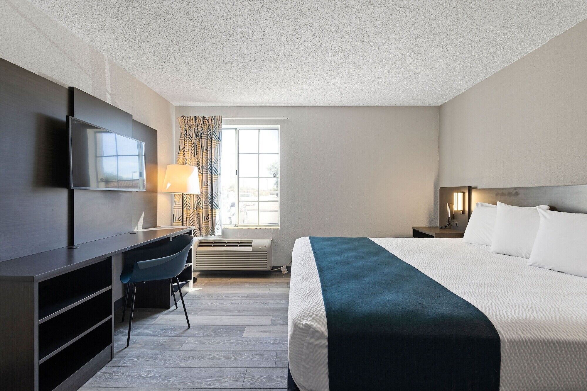 Une chambre d’hôtel comprenant un lit, un bureau, une chaise, une télévision, une fenêtre avec des rideaux et une table de chevet avec une lampe.