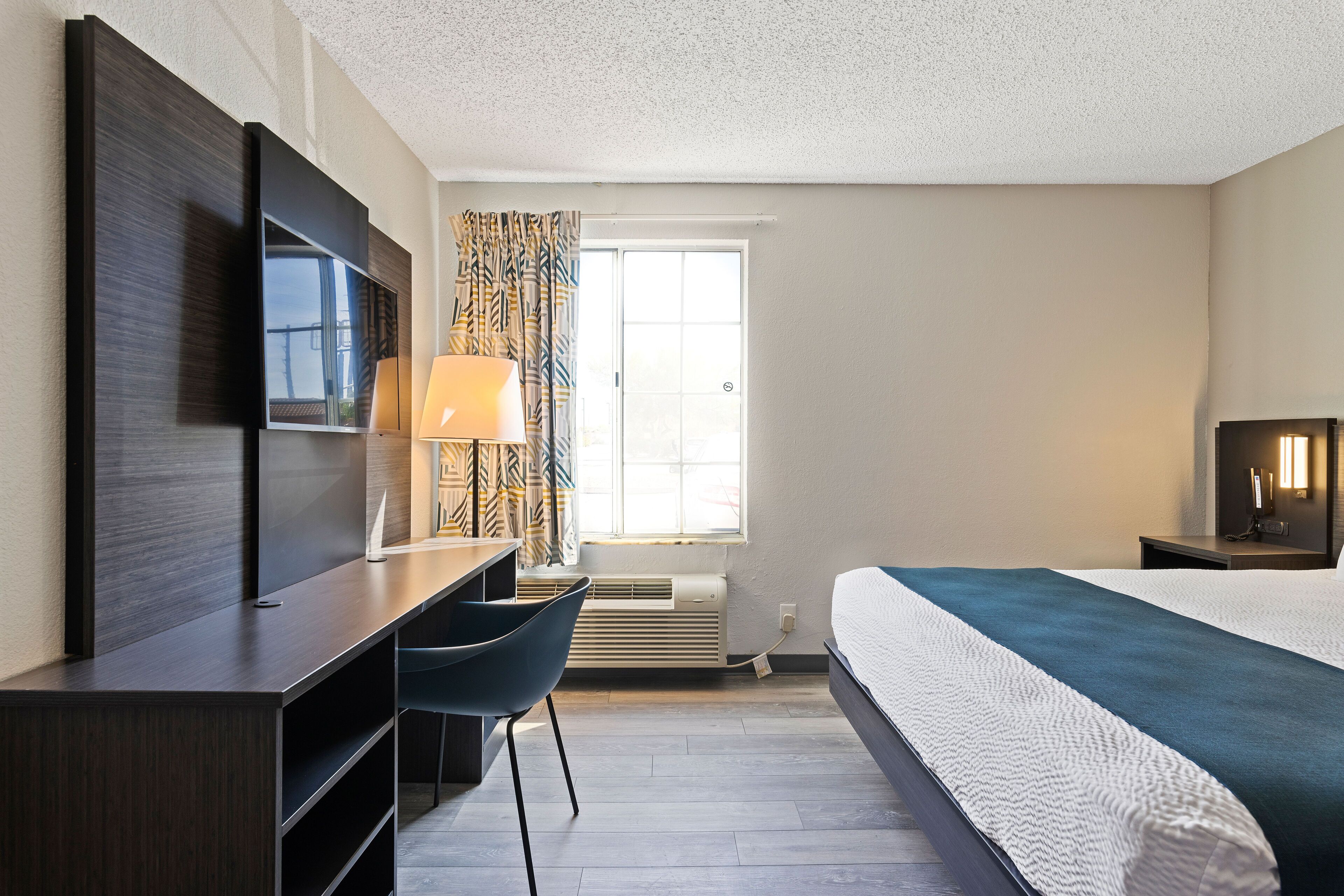 Quarto de hotel com cama, escrivaninha com cadeira, televisão na parede, janela com cortinas e mesa de cabeceira com abajur.