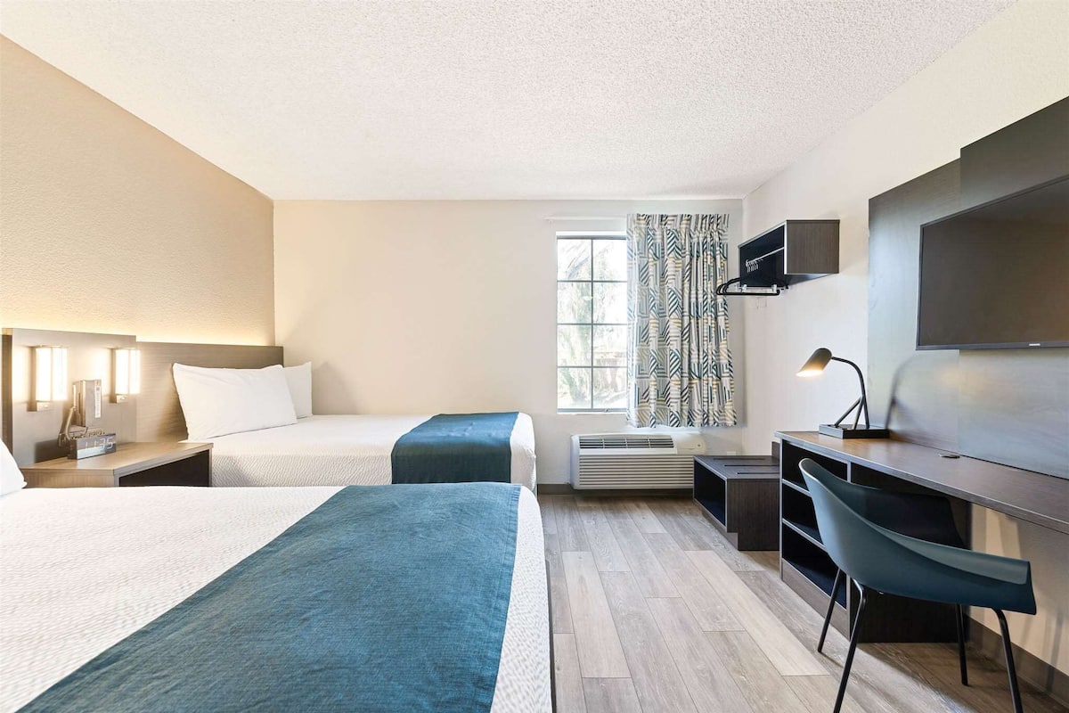 Une chambre d’hôtel avec deux lits, un bureau avec une chaise, une télévision et une fenêtre avec des rideaux.