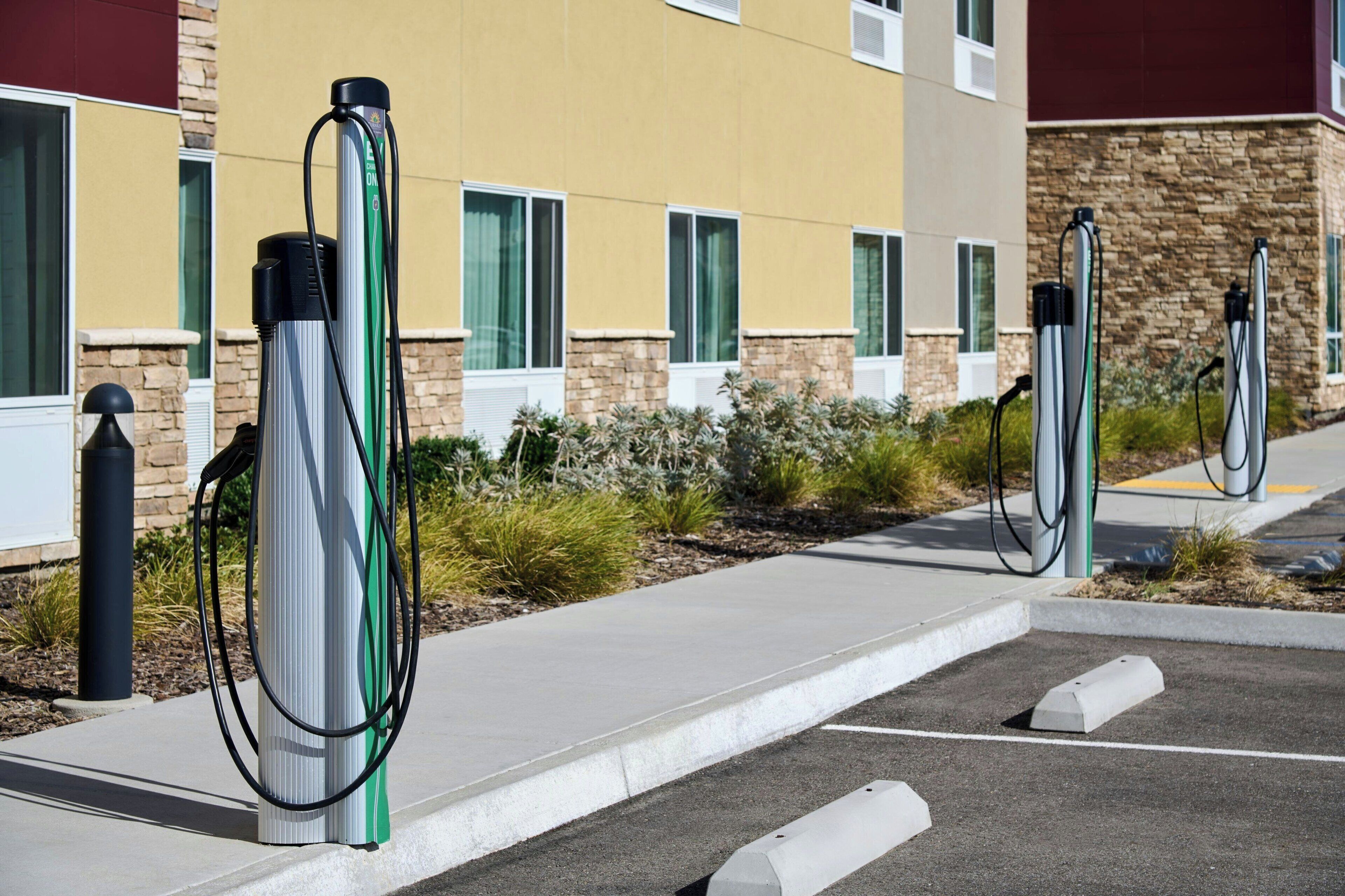 Fileira de postos de carregamento para veículos elétricos em um estacionamento, com um edifício ao fundo.