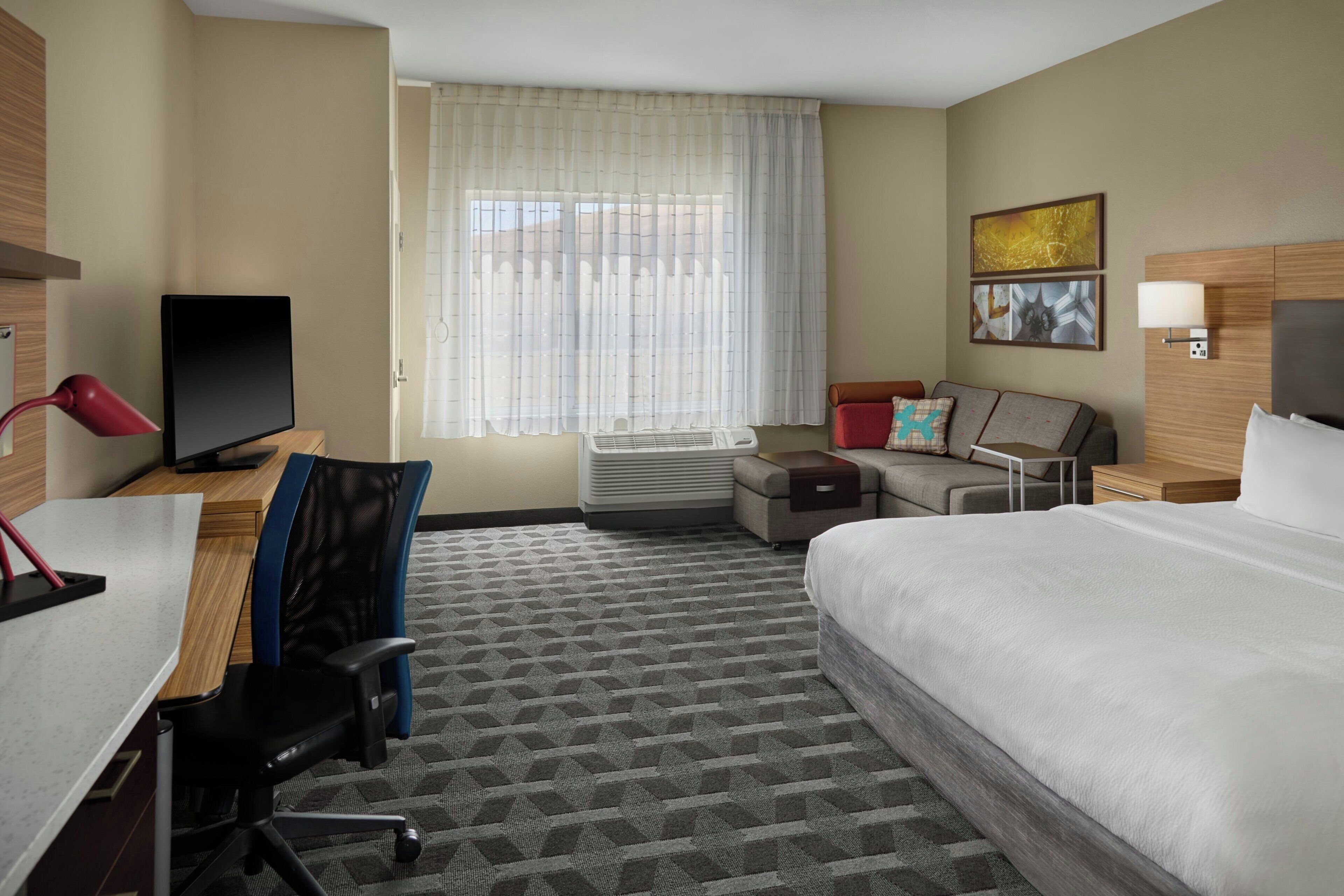 A hotel room with a bed, desk, chair, TV, air conditioning unit, and a window with curtains.