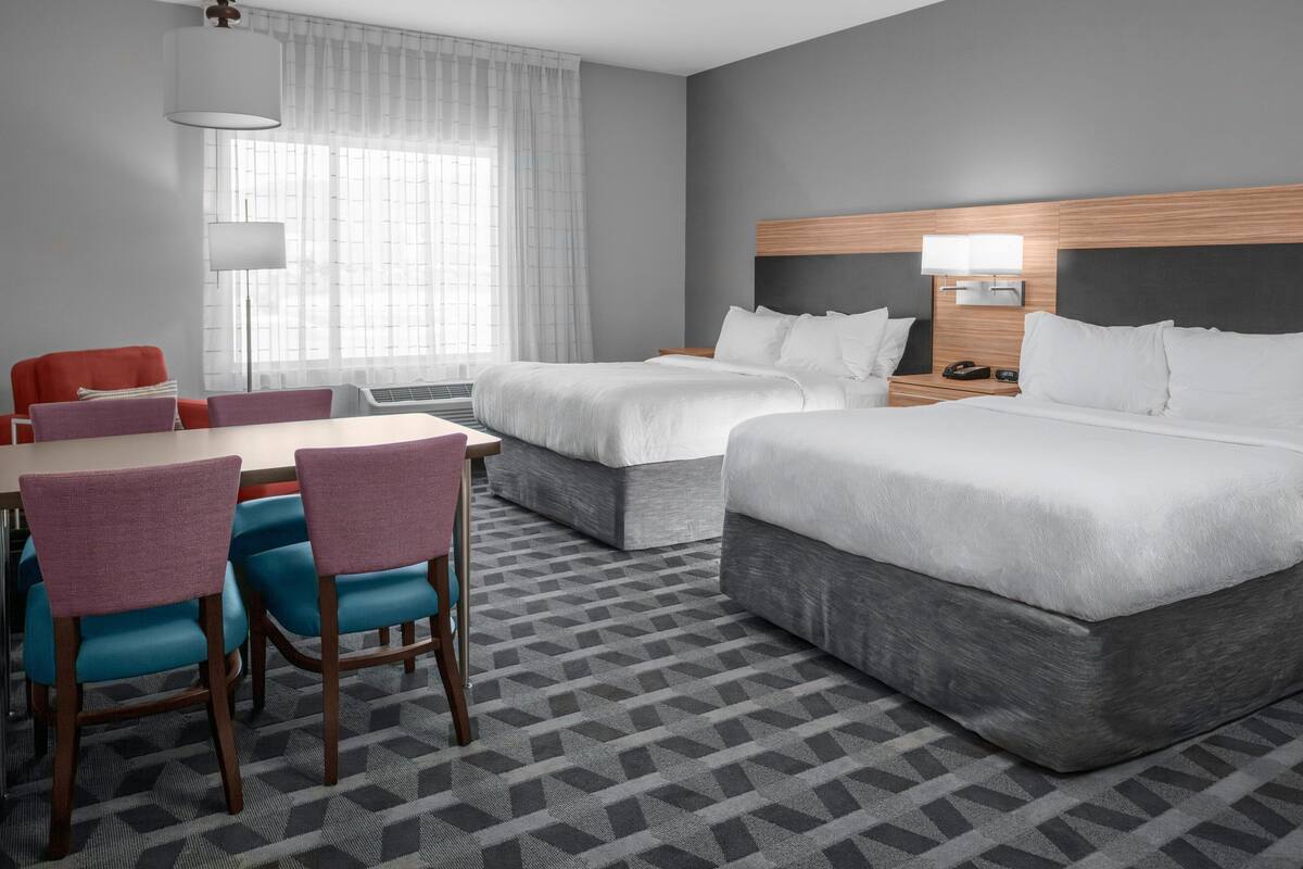 Una habitación de hotel con dos camas, un escritorio con sillas, una lámpara y un ventanal con cortinas.