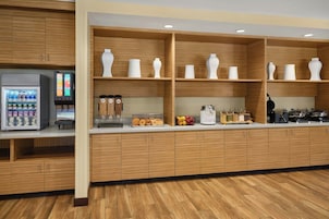 Un área de desayuno autoservicio con dispensador de bebidas refrigeradas, diversas opciones de alimentos y una máquina de café.