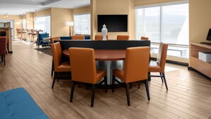 Une chambre d’hôtel moderne avec une table de salle à manger ronde, des chaises orange, un canapé bleu, un téléviseur fixé au mur et de grandes fenêtres offrant une vue dégagée.