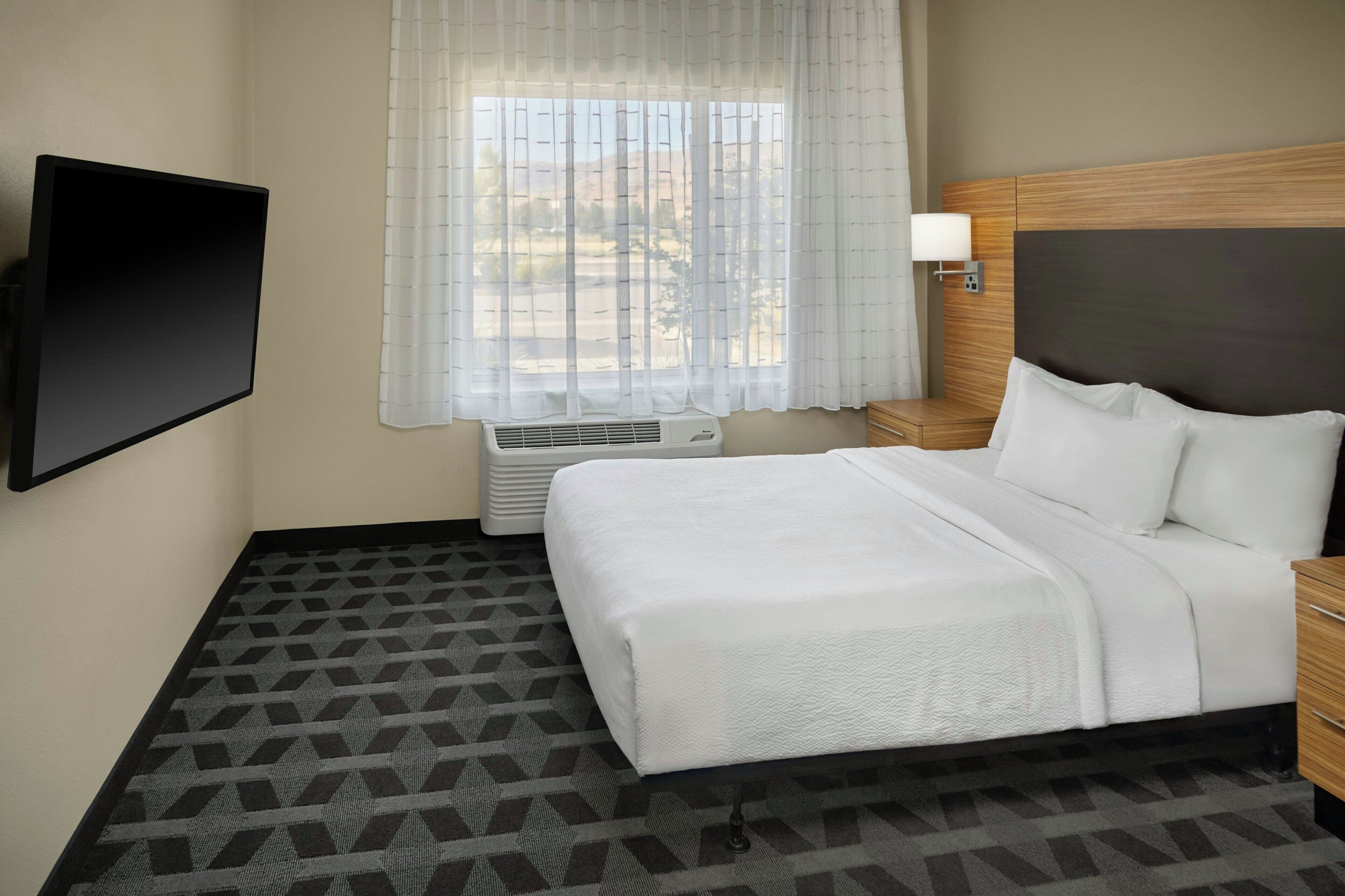 Une chambre d’hôtel comprenant un lit, un téléviseur à écran plat fixé au mur, une fenêtre avec des rideaux légers, un appareil de climatisation et une moquette à motifs.