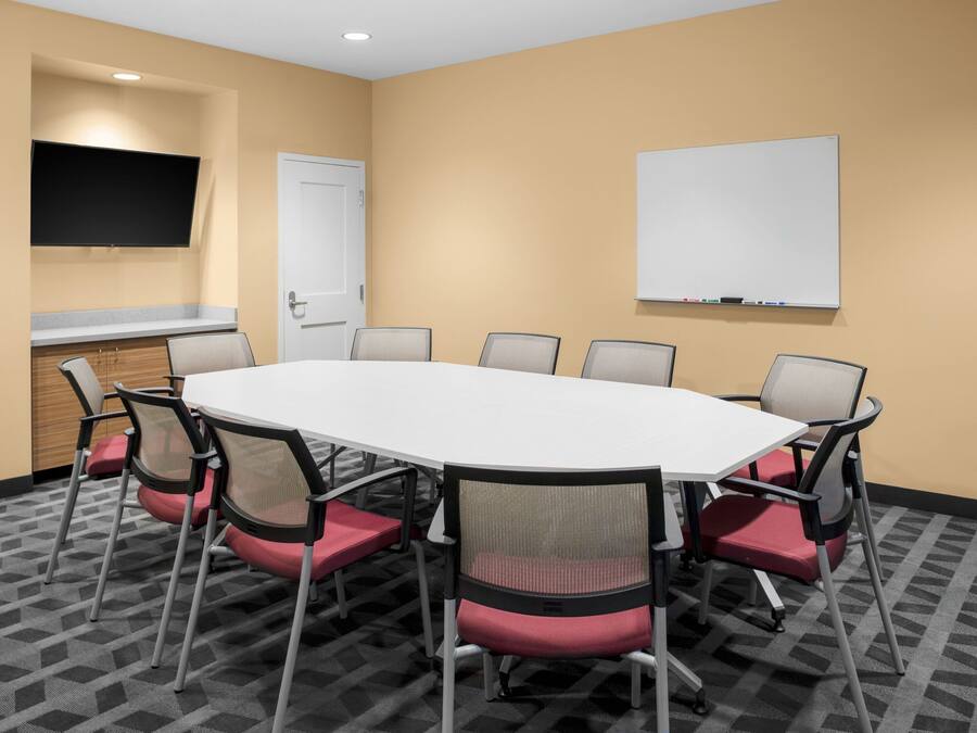 A conference room with a white table, red and black chairs, a whiteboard, a flat-screen TV, and a door.