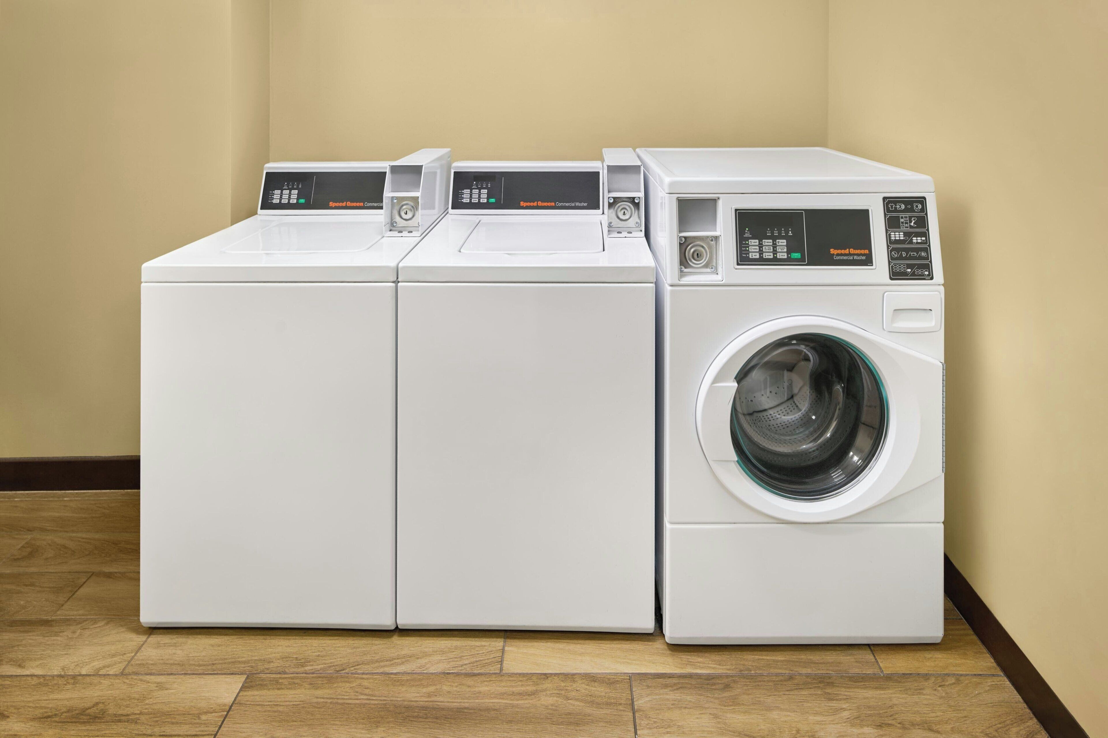 A row of three white commercial washers with control panels, placed against a beige wall in a corner.
