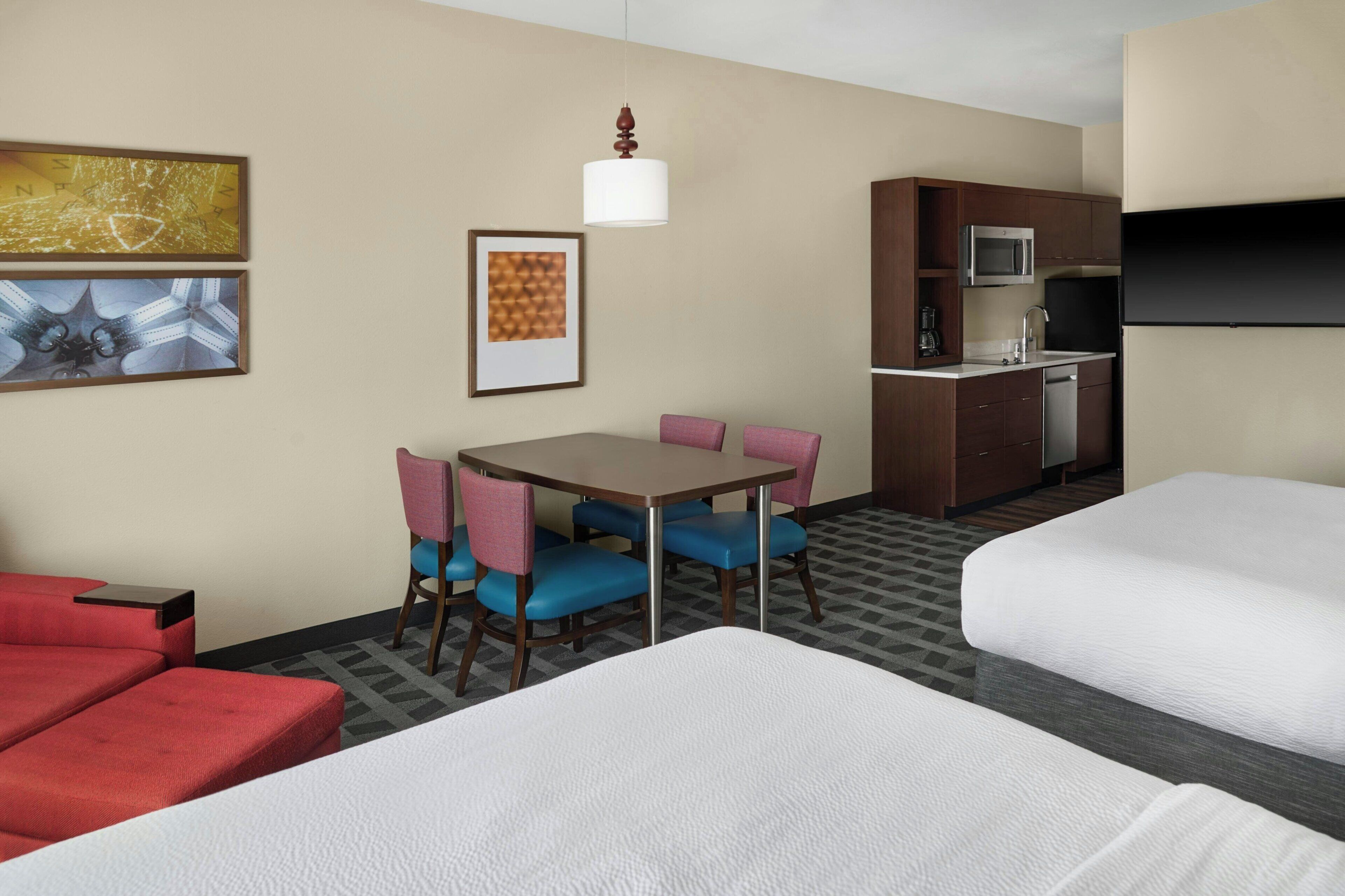 Habitación de hotel con dos camas, una mesa de comedor con sillas, una kitchenette y obras de arte en las paredes.