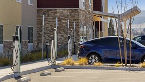 Un parking équipé de bornes de recharge pour véhicules électriques, une voiture bleue et un bâtiment dont la façade est en pierre et en bois.