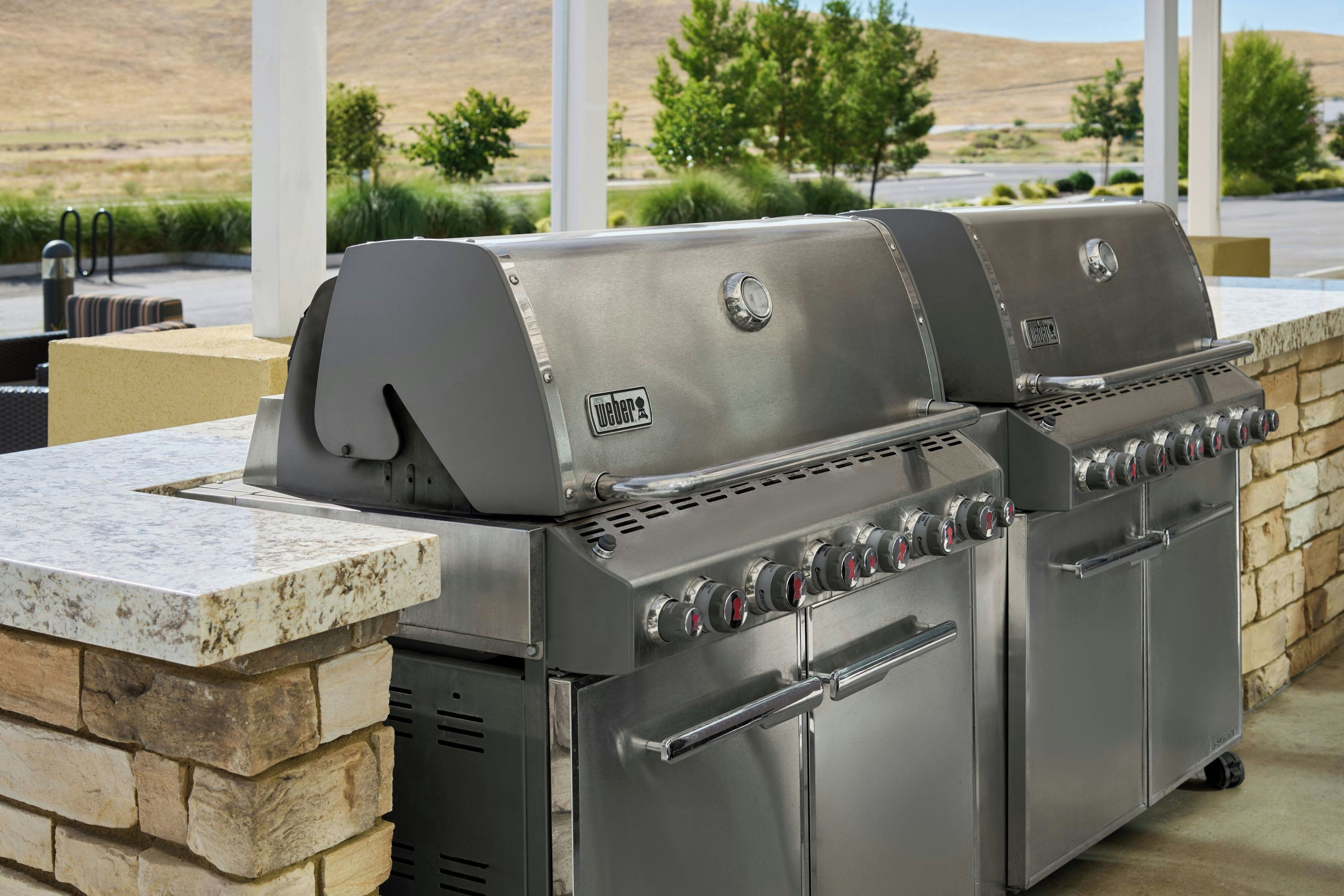 Una parrilla de gas Weber con múltiples quemadores y un armario lateral, frente a una pared de piedra con vistas a una zona ajardinada.