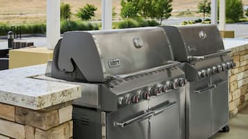Una parrilla de gas Weber con múltiples quemadores y un armario lateral, frente a una pared de piedra con vistas a una zona ajardinada.