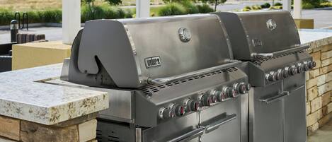 Una parrilla de gas Weber con múltiples quemadores y un armario lateral, frente a una pared de piedra con vistas a una zona ajardinada.