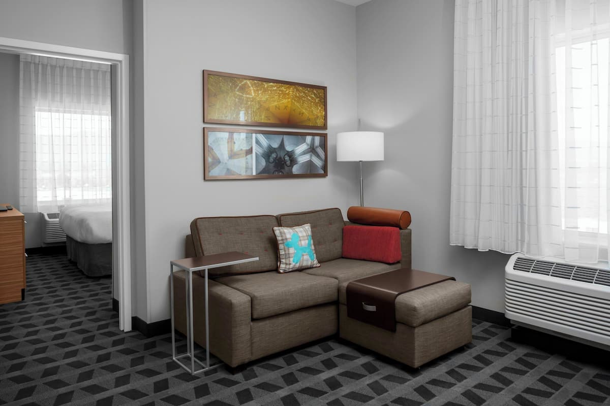 Una habitación de hotel moderna con sofá modular, una mesita, una lámpara y un amplio ventanal con cortinas.
