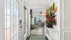 A hallway with a white door, a glass door with a floral pattern, a white console table with a floral arrangement, and a bookshelf in the background. - Luxury Beach Estate Ocean view, Pool, 360° Deck&Beach Access 20%off last minute  (Surfside Beach)
