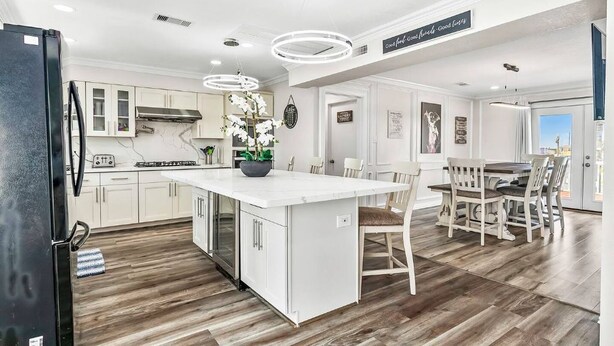 A modern kitchen with white cabinetry, a large island, and a dining area with wooden chairs. - Luxury Beach Estate Ocean view, Pool, 360° Deck&Beach Access 20%off last minute  (Surfside Beach)