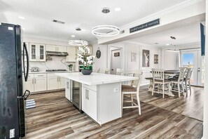 A modern kitchen with white cabinetry, a large island, and a dining area with wooden chairs. - Luxury Beach Estate Ocean view, Pool, 360° Deck&Beach Access 20%off last minute  (Surfside Beach)