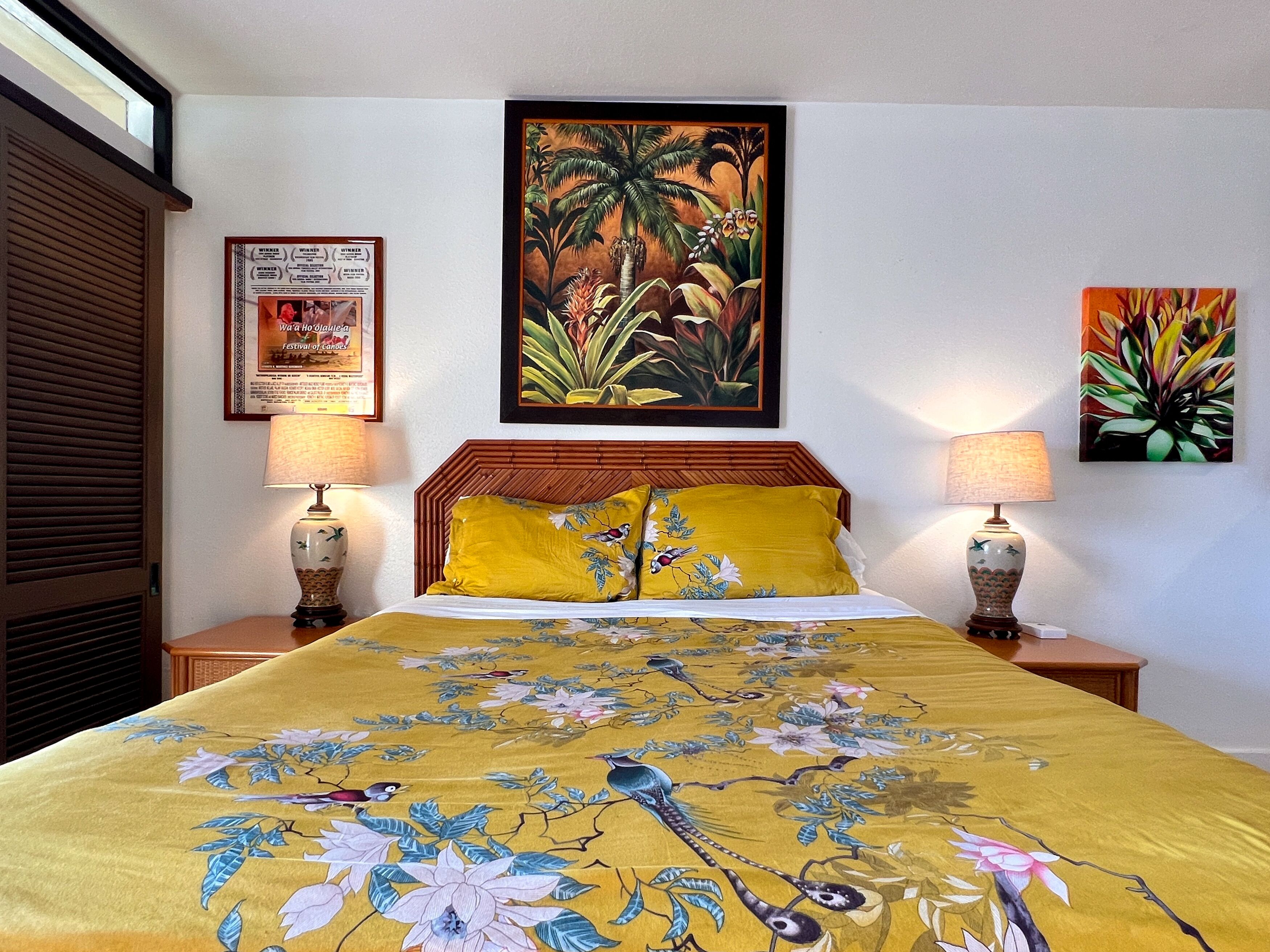 Una habitación de hotel con una cama, dos mesitas de noche, dos lámparas, un cuadro con motivos tropicales y un cartel enmarcado en la pared.