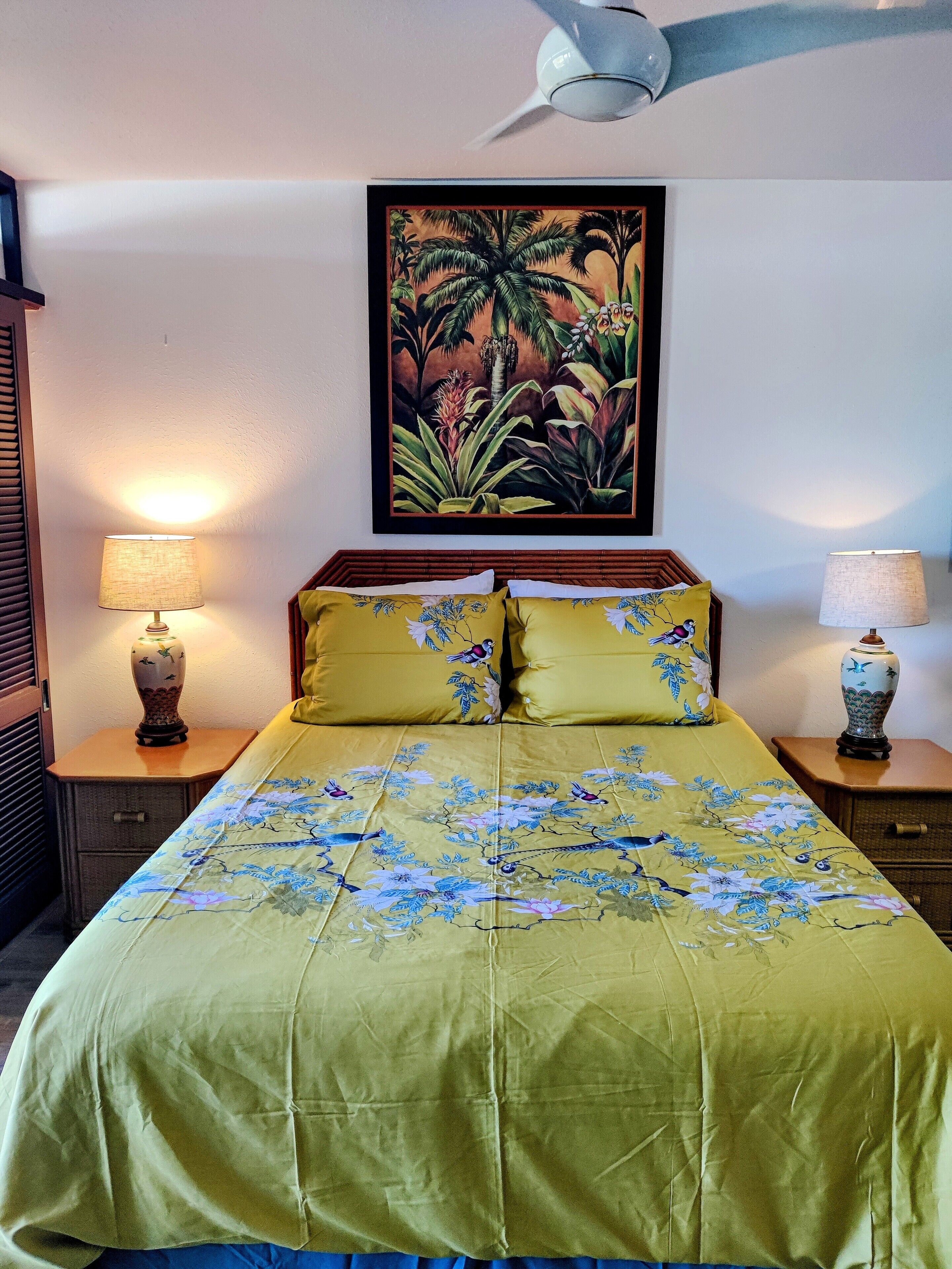 Una cama bien tendida con ropa de cama floral, dos lámparas de noche, un ventilador de techo y un cuadro con motivos tropicales sobre la cama.