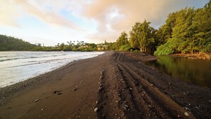 Une plage avec un sentier sablonneux menant à l’eau, entourée d’arbres et bordée d’un bâtiment situé au loin.