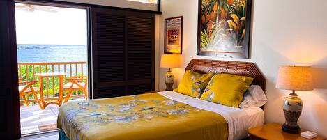 A bedroom with a bed, a nightstand, a lamp, a painting, and a view of the ocean through a sliding door.