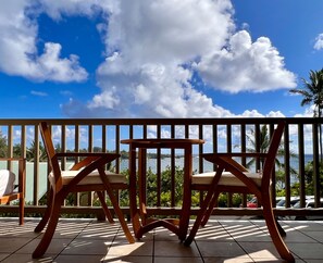 A wooden deck with two chairs and a small table, overlooking a body of water and palm trees. - Oceanview Condo in Hana-Let the ocean lull you to sleep (Hana)