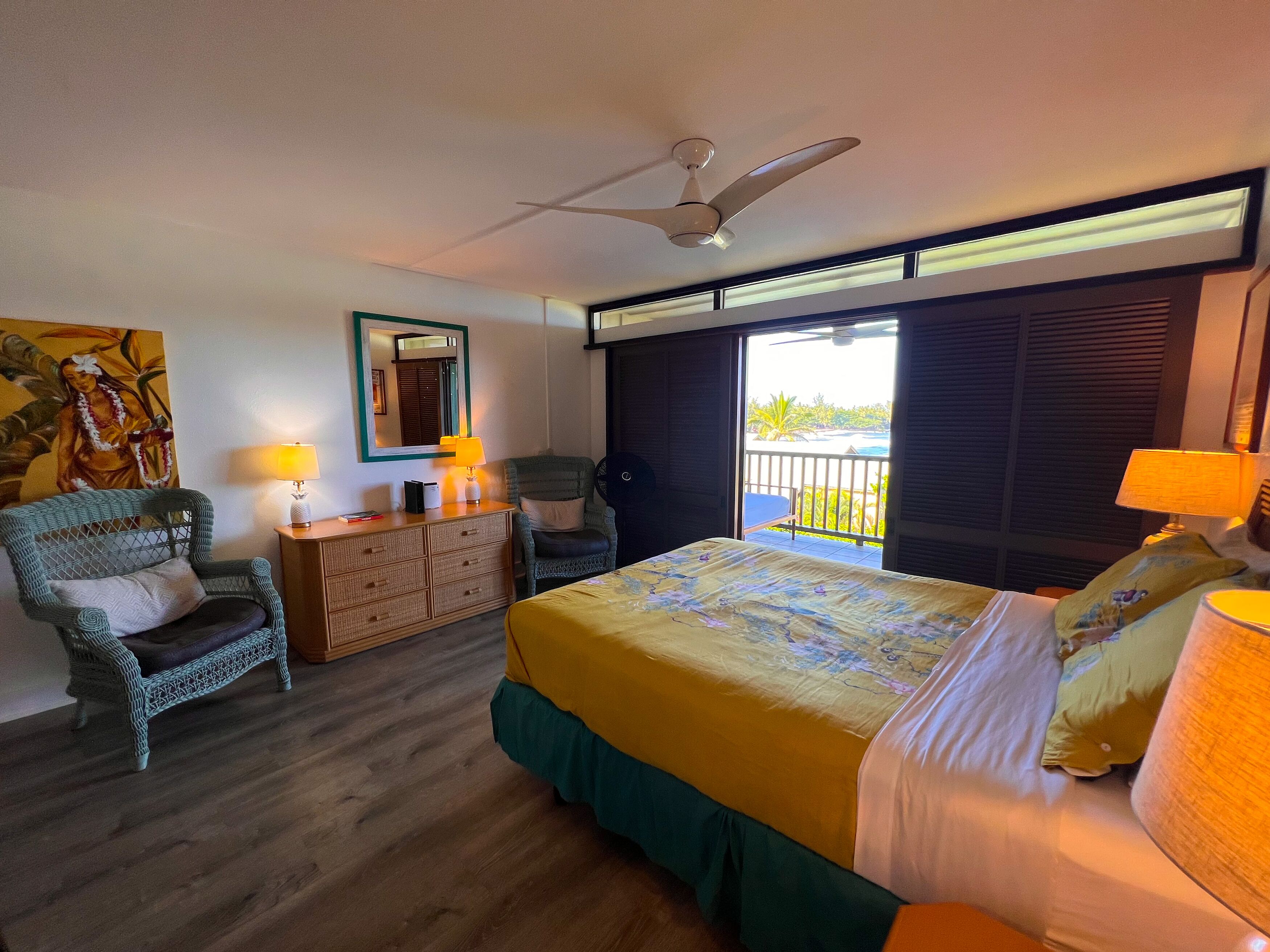 A hotel room with a bed, a desk, a chair, a ceiling fan, and a view of the outdoors.