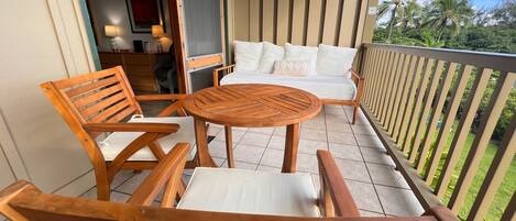 A wooden deck with a round table and two chairs, a sofa with white cushions, and a wooden railing overlooking a garden with palm trees.