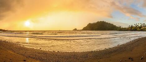 Ein Strand mit Wellen, einem Sonnenuntergang und einer bewaldeten Hügelkette im Hintergrund.