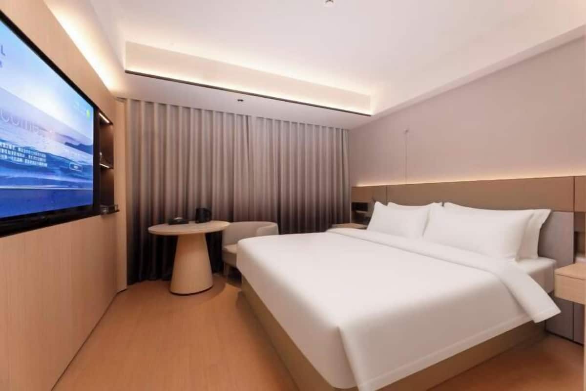 A modern hotel room with a large bed, a small round table, a TV mounted on the wall, and a curtain-covered window.