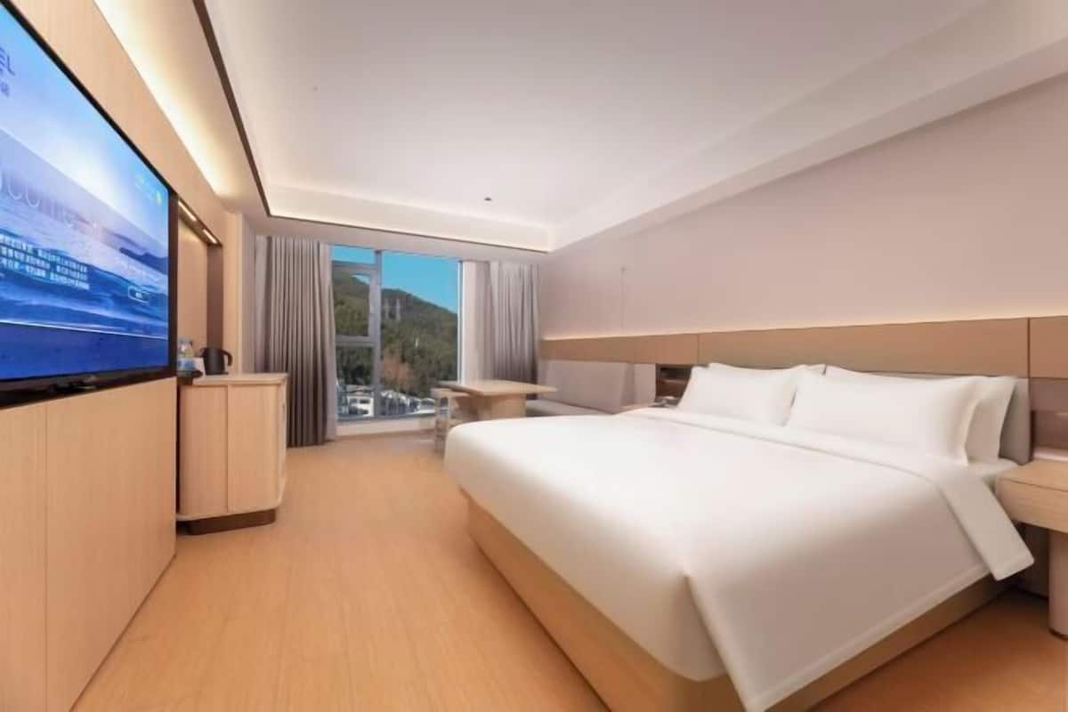 A modern hotel room with a large bed, a flat-screen TV, a desk, and a view of a mountain.