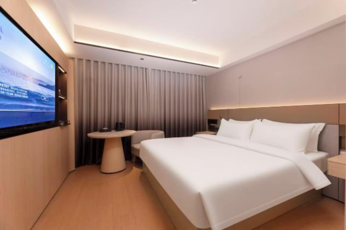 A modern hotel room with a large bed, a small round table, a TV mounted on the wall, and a curtain-covered window.