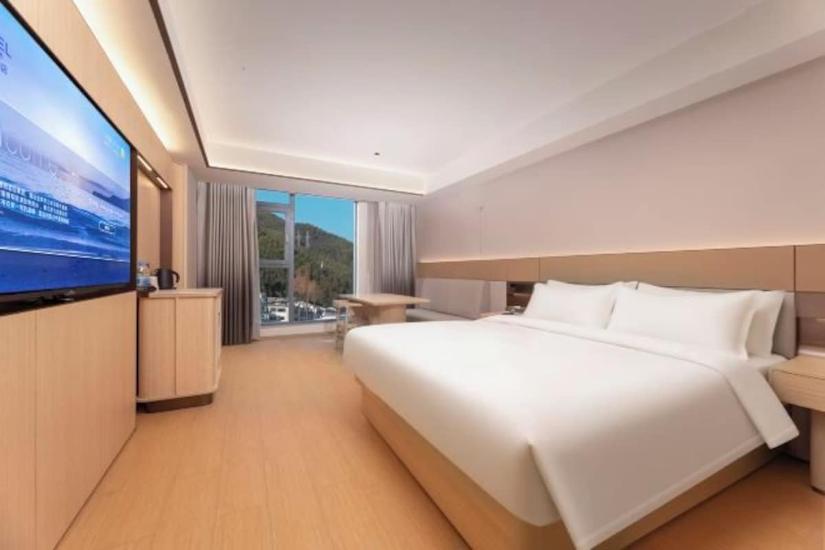 A modern hotel room with a large bed, a flat-screen TV, a desk, and a view of a mountain.
