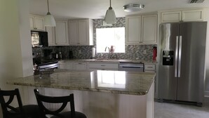 A kitchen with white cabinets, a stainless steel refrigerator, a marble island, and two black chairs. - Riverside Retreat (Dunnellon)