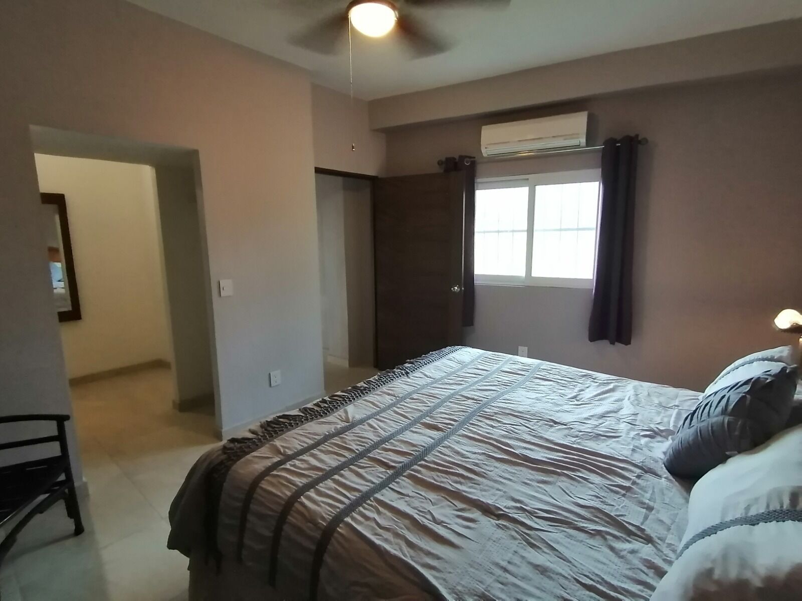A bedroom with a bed, a ceiling fan, an air conditioning unit, a window with curtains, and a door leading to another room.