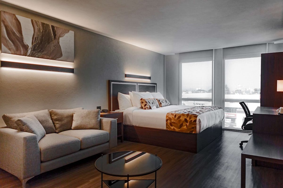 Una habitación de hotel moderna con cama, sofá, una mesita y un ventanal con vistas a la ciudad.