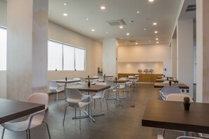 Uma ampla área de jantar com várias mesas e cadeiras, um balcão longo com vitrine e grandes janelas que proporcionam iluminação natural.