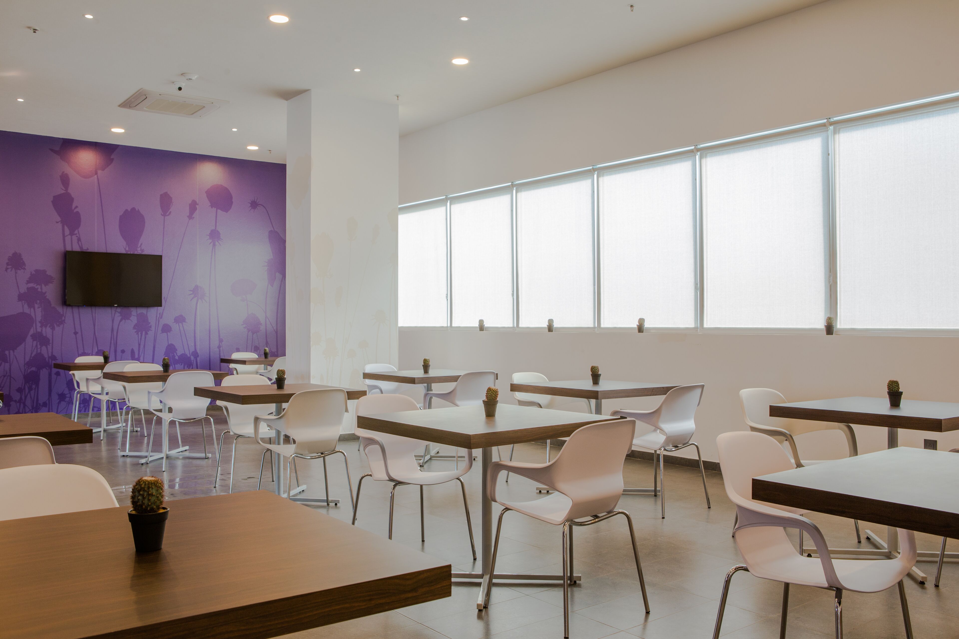 A modern dining area with white chairs and tables, a flat-screen TV mounted on the wall, and large windows providing natural light.