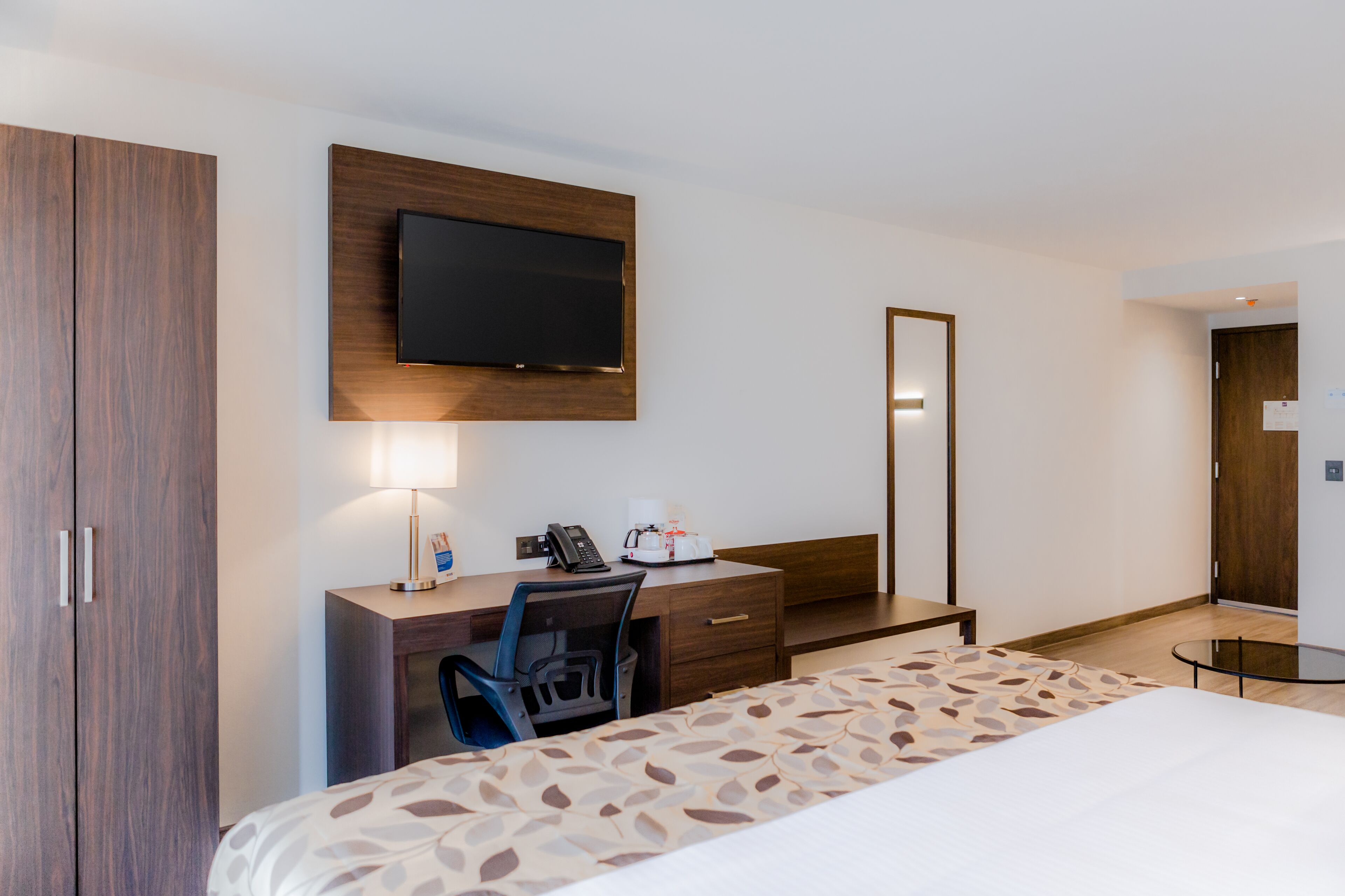 A hotel room with a bed, desk, chair, and a flat-screen TV mounted on the wall.