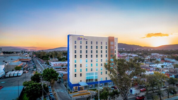 Um hotel Sleep Inn com design moderno, destacando um esquema de cores azul e branco, situado em uma paisagem urbana com vista para o pôr do sol e edifícios ao redor.