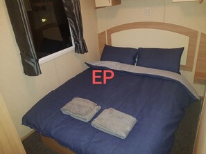 A single bed with a blue bedspread and pillows, a window with curtains, and a wooden headboard. - Lovely 3-bed Caravan in Towyn (Abergele)